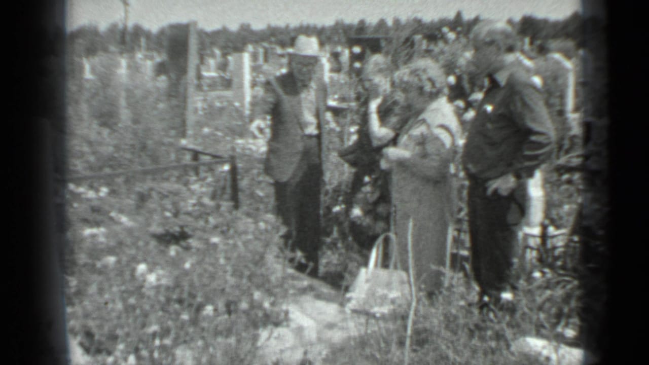 Adults stand among graves and overgrown grass in a cemetery.