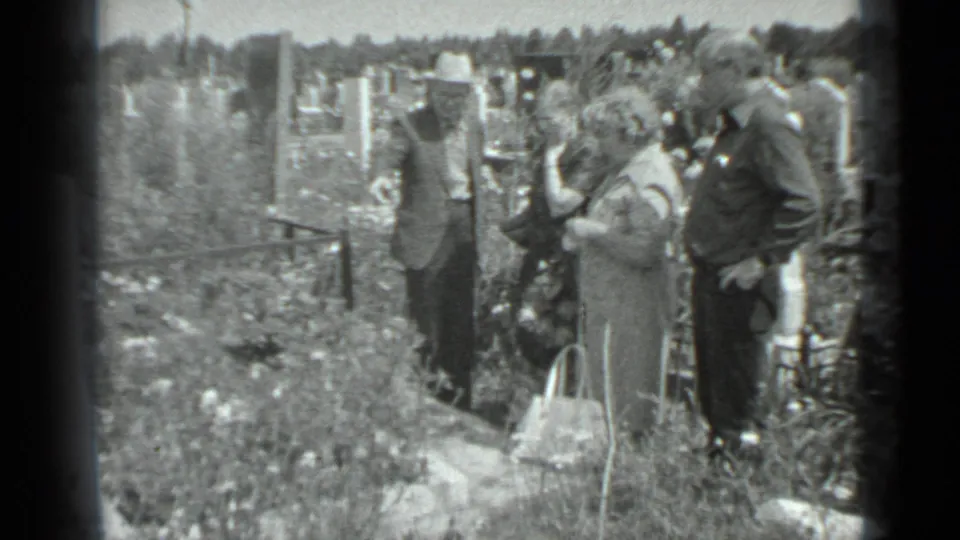In Moscow, 1983, adults gather quietly beside a gravesite, with hats, handbags, grave markers, and tall grass visible.