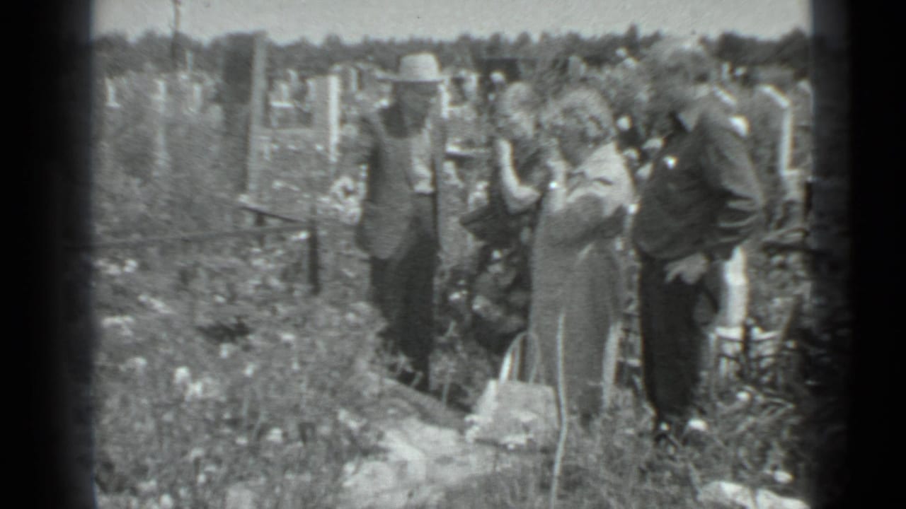 Adults stand near grave markers while a woman holds a handbag.