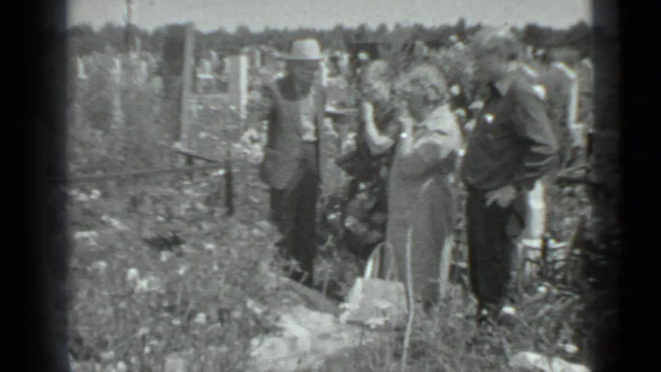 A small group of adults stands in a Moscow cemetery, with a woman and handbag in the foreground and grave markers behind.