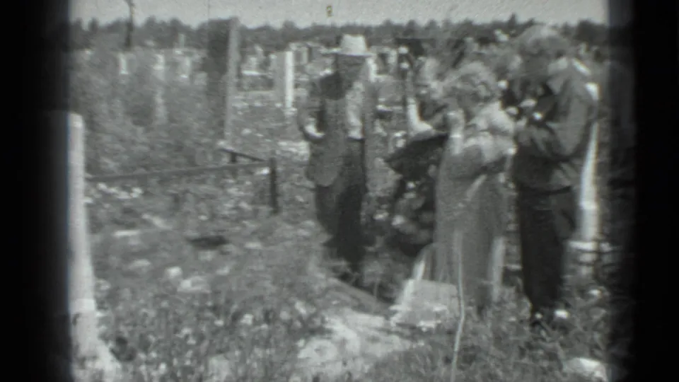 Adults in a grassy Moscow cemetery appear to adjust their positions near a grave, with hats, bags, and markers visible.