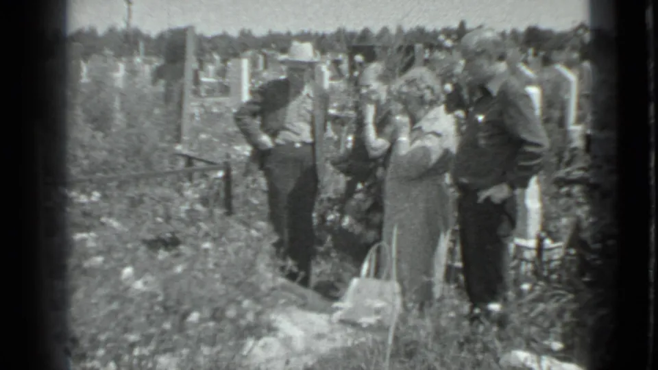 A Moscow cemetery visit shows adults standing close together near a grave, with tall grass, markers, and a handbag in view.