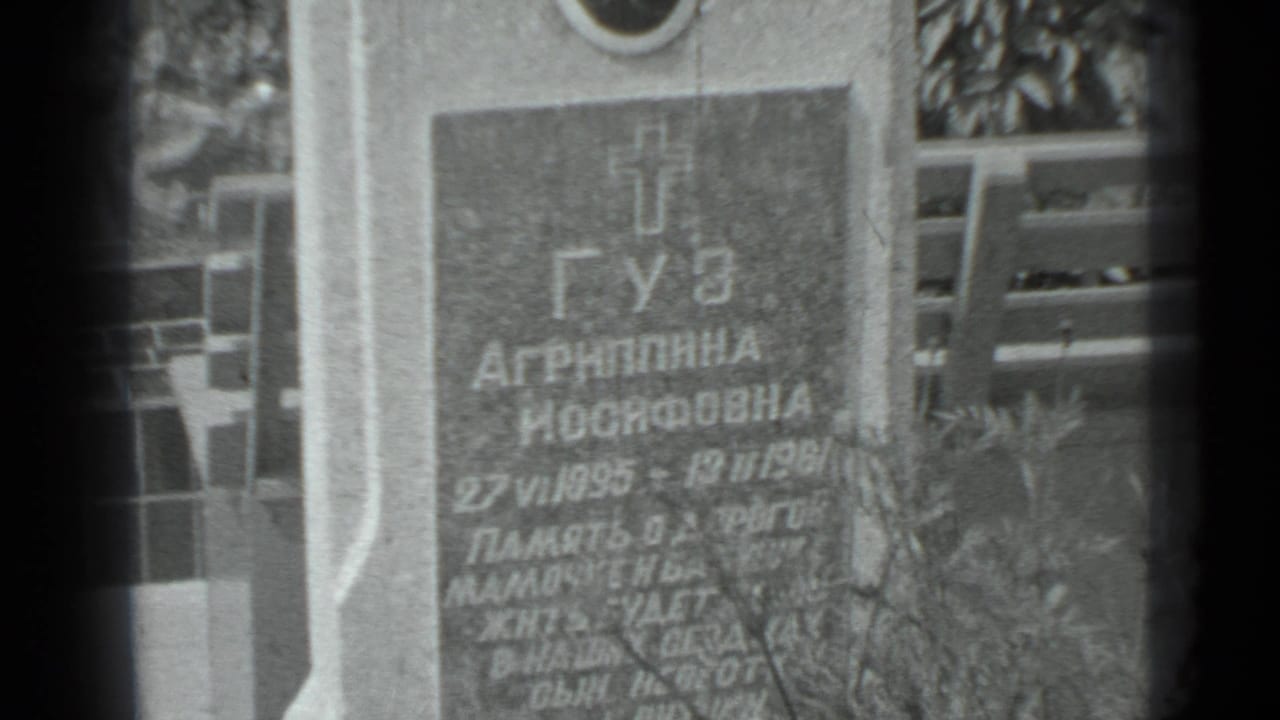 Cyrillic gravestone seen closer, with inscription, portrait area, and plants in front.