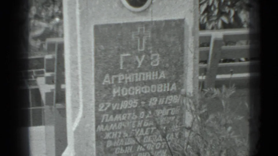 Closer view of the Moscow cemetery gravestone, with Cyrillic lettering, portrait plaque area, cross motif, and overgrown plants.