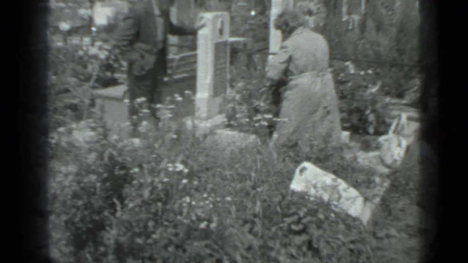 Adults in a Moscow cemetery, supported by metadata, stand quietly near gravestones, flowers, and dense plantings during a memorial visit.