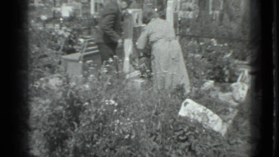 Two adults stand beside a grave in a Moscow cemetery, with flowers, plants, gravestones, and soft 8mm home-movie framing.