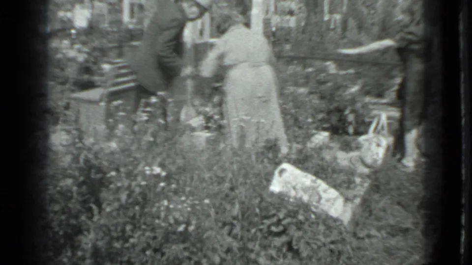 In 1983 Moscow home-movie footage, adults appear to tend an overgrown cemetery plot with grass, plants, and grave markers around them.