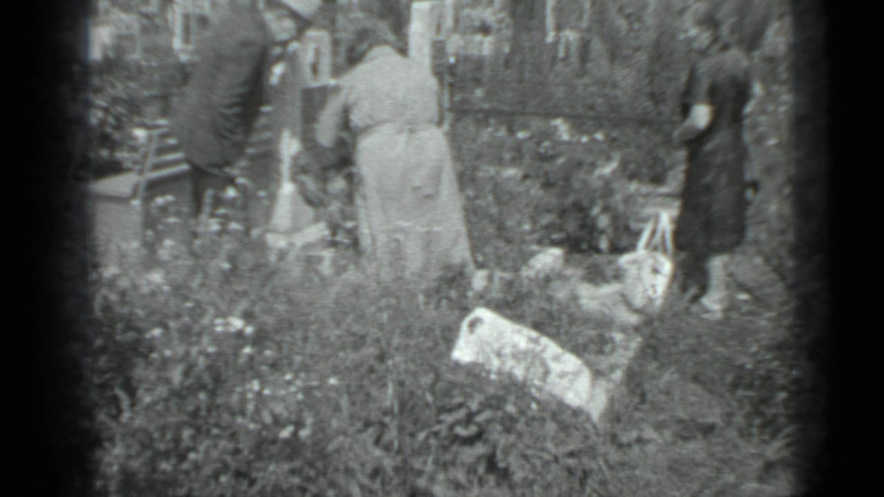 Adults are seen from behind near tall grass, flowers, and grave markers in a cemetery-like yard.