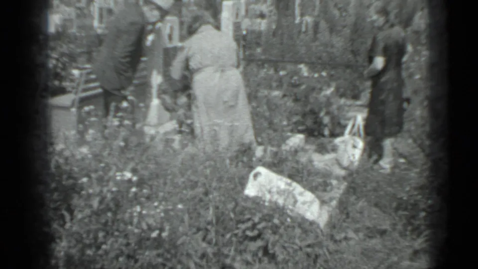 Adults in 1983 Moscow appear from behind while standing among flowers, grass, and grave markers in a cemetery-like setting.