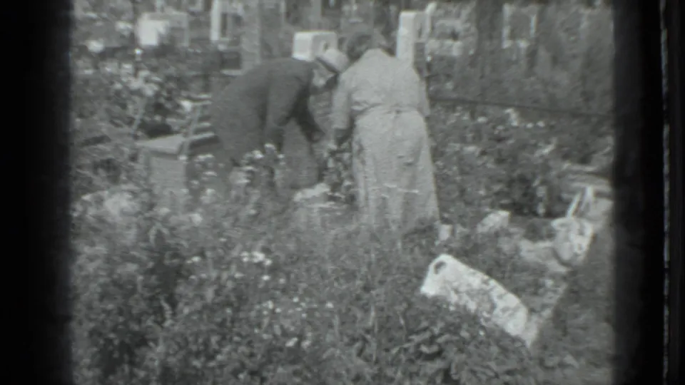 Two adults in 1983 Moscow bend over a cemetery plot, with a pale grave marker, tall grass, flowers, and a bag visible nearby.