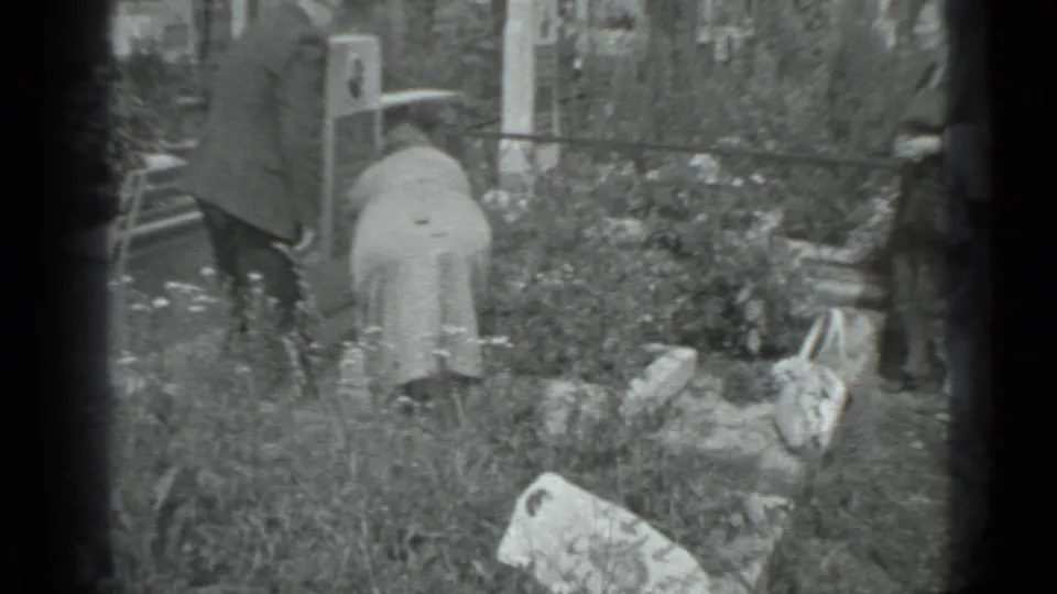 The older man and woman continue tending a Moscow garden, surrounded by wildflowers, tall grass, and household garden items.