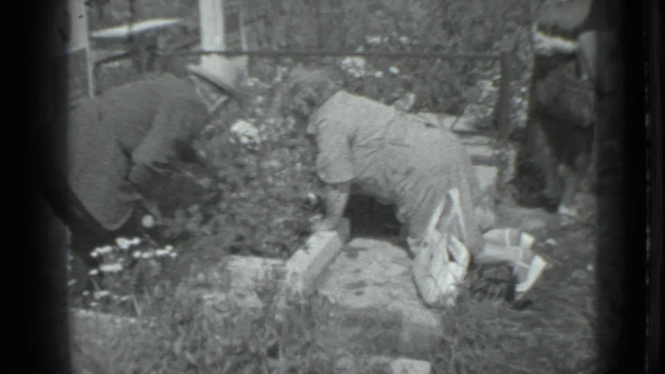 The couple kneels and bends over the garden bed, with soft black-and-white 8mm grain and tall flowers filling the frame.