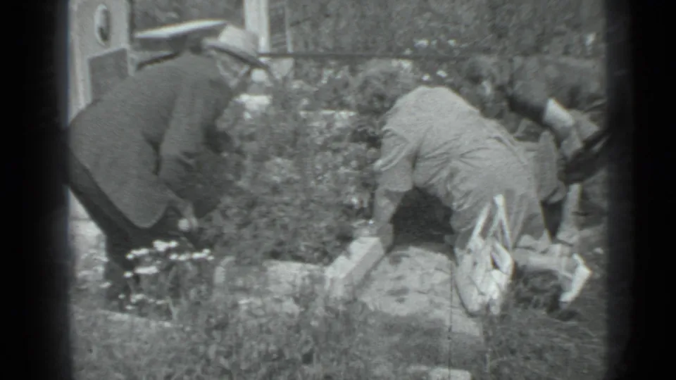 In 1983 Moscow home-movie footage, adults bend over a grave area while flowers and low markers sit among cemetery greenery.