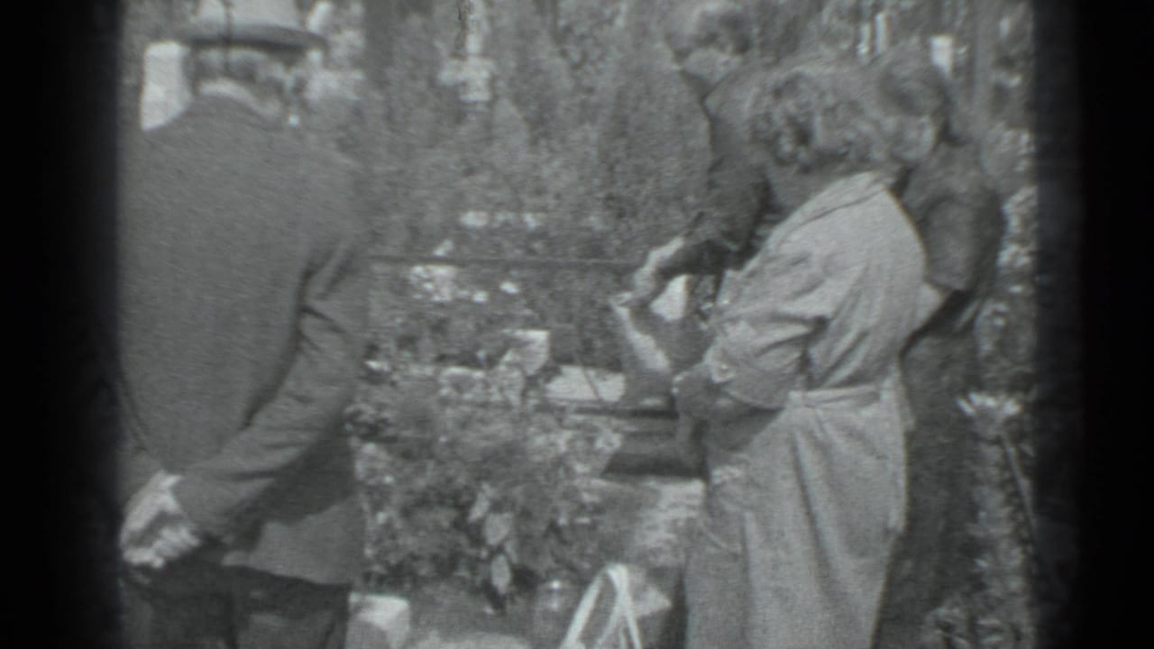 Man in a suit and woman in a light dress stand near a grave.