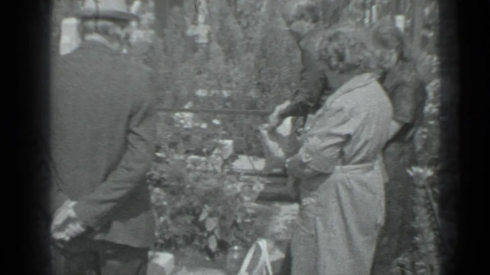 A man in a dark suit and hat stands with his hands behind his back as a woman in a light dress watches near a Moscow cemetery grave.