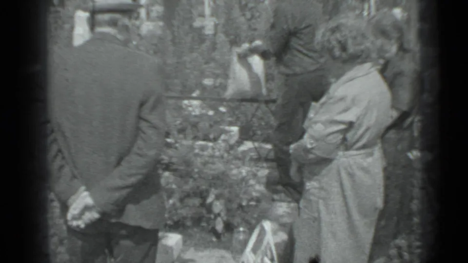The home-movie frame shows adults gathered beside planted flowers and grave markers in a Moscow cemetery, with the camera held close.
