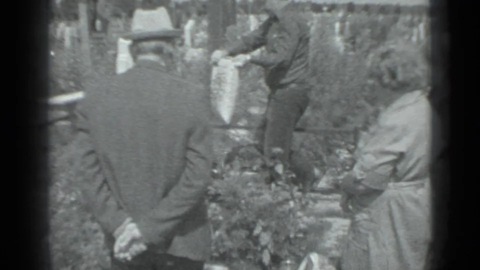 A visitor reaches toward a grave area as a man in a hat and a woman in a light dress stand nearby in the cemetery.