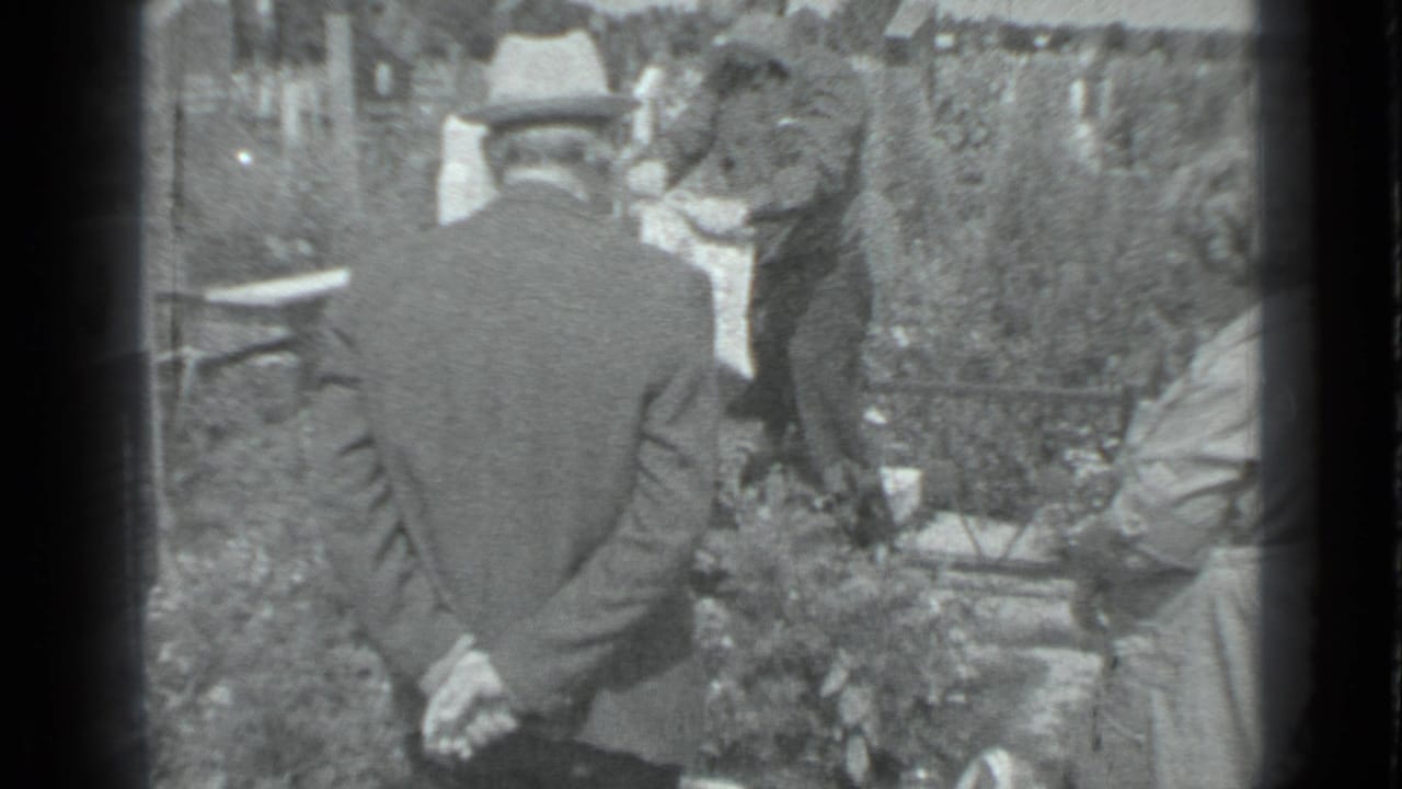 Mourners stand near a grave behind a man in a fedora with his hands clasped.