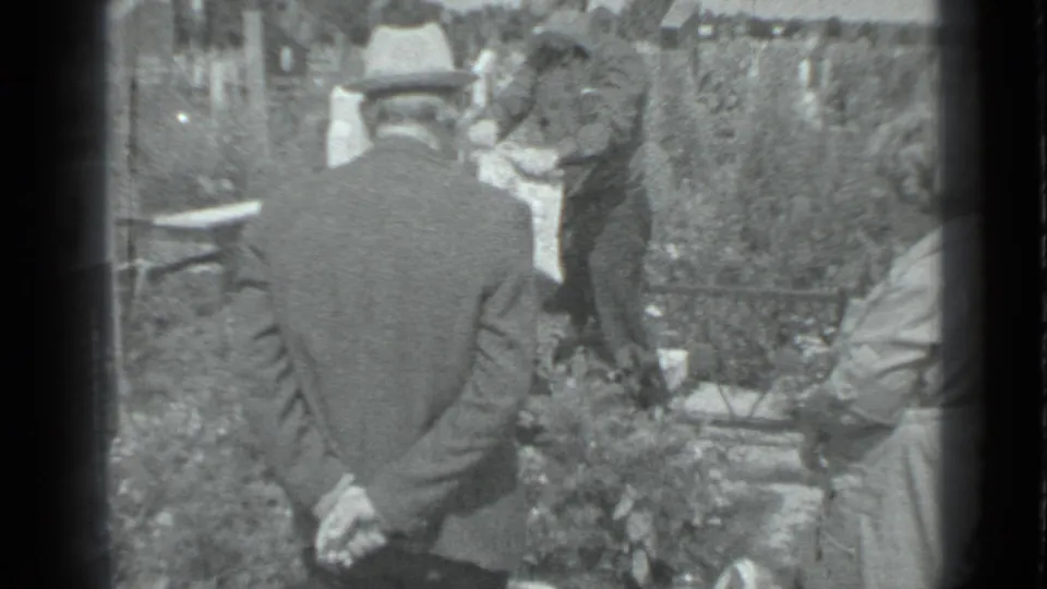 1983 Moscow cemetery footage showing a man in a fedora from behind as mourners gather by flowers and grave markers.