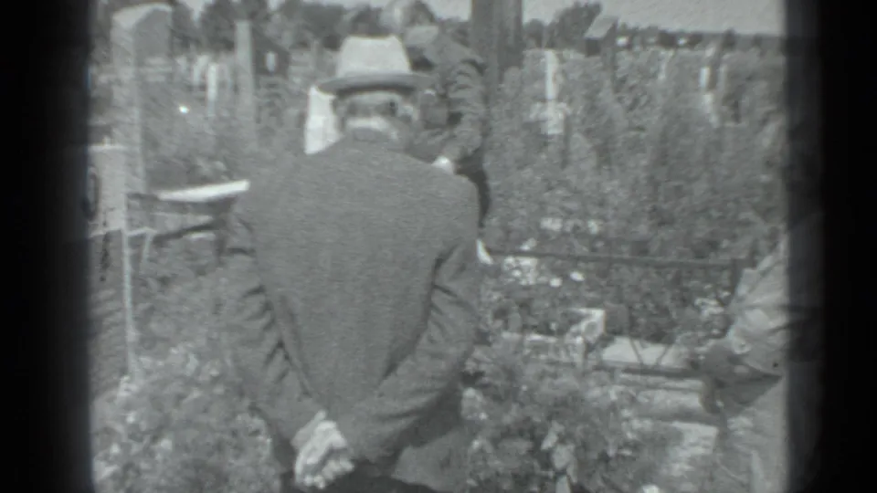 In the Moscow cemetery scene, a suited man faces the graveside while another mourner is partly visible at the right edge.