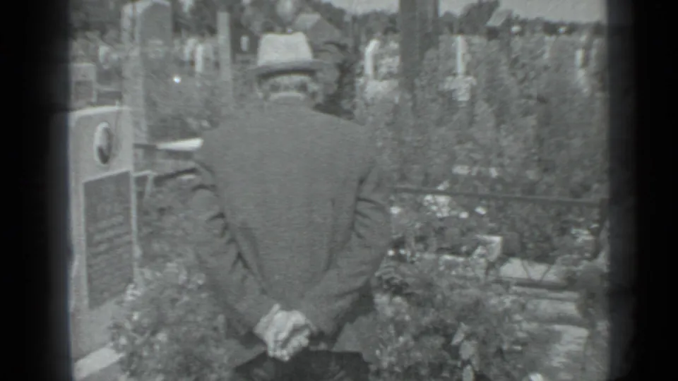 The handheld 8mm frame shows the mourner from behind beside a grave marker, flowers, fencing, and trees in Moscow.