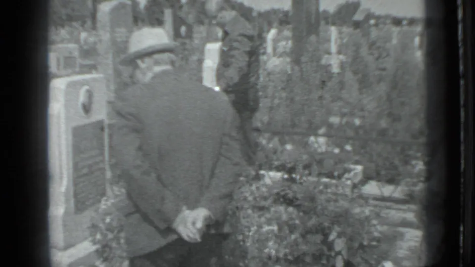The closing frame keeps the man in the foreground, with Moscow cemetery flowers, grave markers, and a partial mourner nearby.