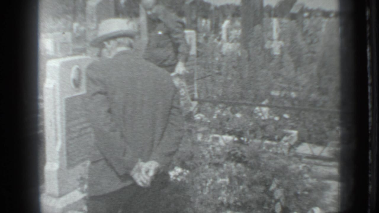 In 1983 Moscow home-movie footage, a man in a cap and jacket stands beside a cemetery gravestone with plantings and markers nearby.