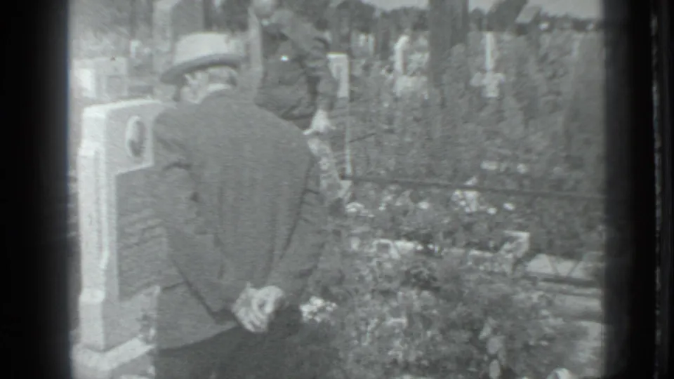 In 1983 Moscow home-movie footage, a man in a cap and jacket stands beside a cemetery gravestone with plantings and markers nearby.