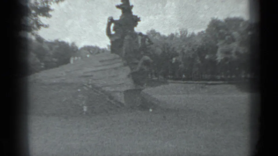 The handheld 8mm view frames a large Moscow monument and sloped base across a lawn, with trees and park landscaping behind it.