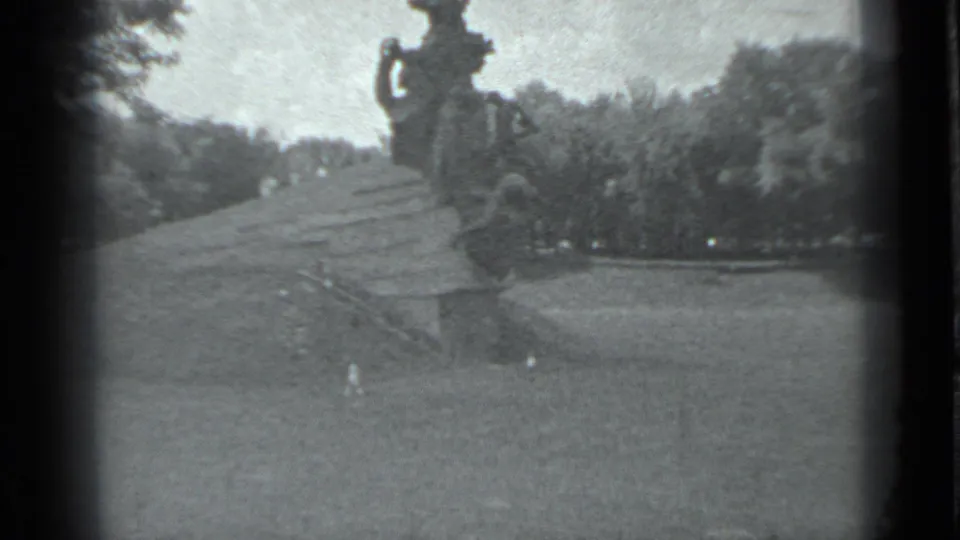 A wide 1983 Moscow home-movie frame shows the outdoor monument, broad base, lawn, and tree-lined background in soft black-and-white footage.