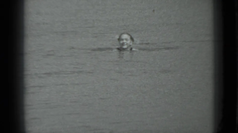 A young woman smiles toward the camera while swimming, framed by grainy black-and-white 8mm film borders.