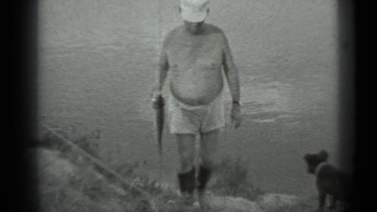Shirtless man in shorts and boots stands near water holding a fish as a small dog watches.