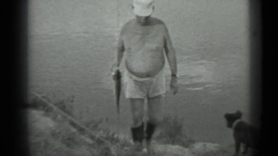In 1983 Moscow home-movie footage, a shirtless man in shorts and boots stands on a riverbank holding a fish while a small dog looks on.