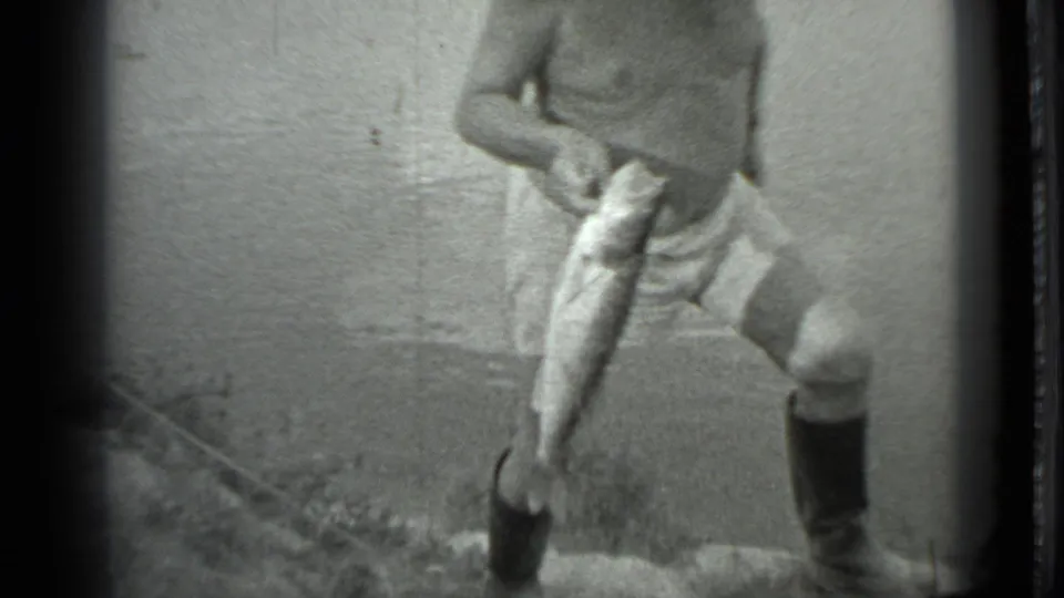 A closer 8mm frame shows the man lifting one boot onto the muddy bank while holding the freshly caught fish in front of him.