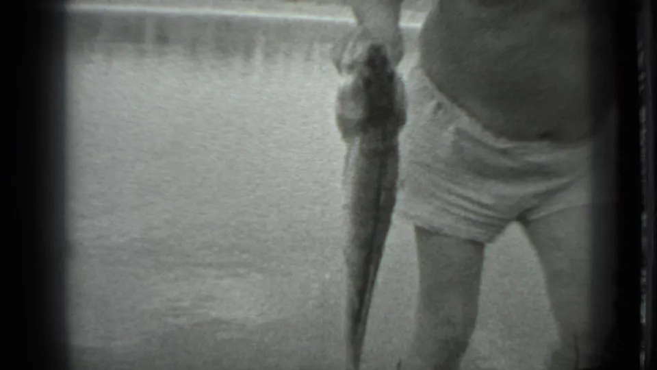 In 1983 Moscow home-movie footage, a man in swim trunks and a brimmed hat holds up a long fish near a lake.