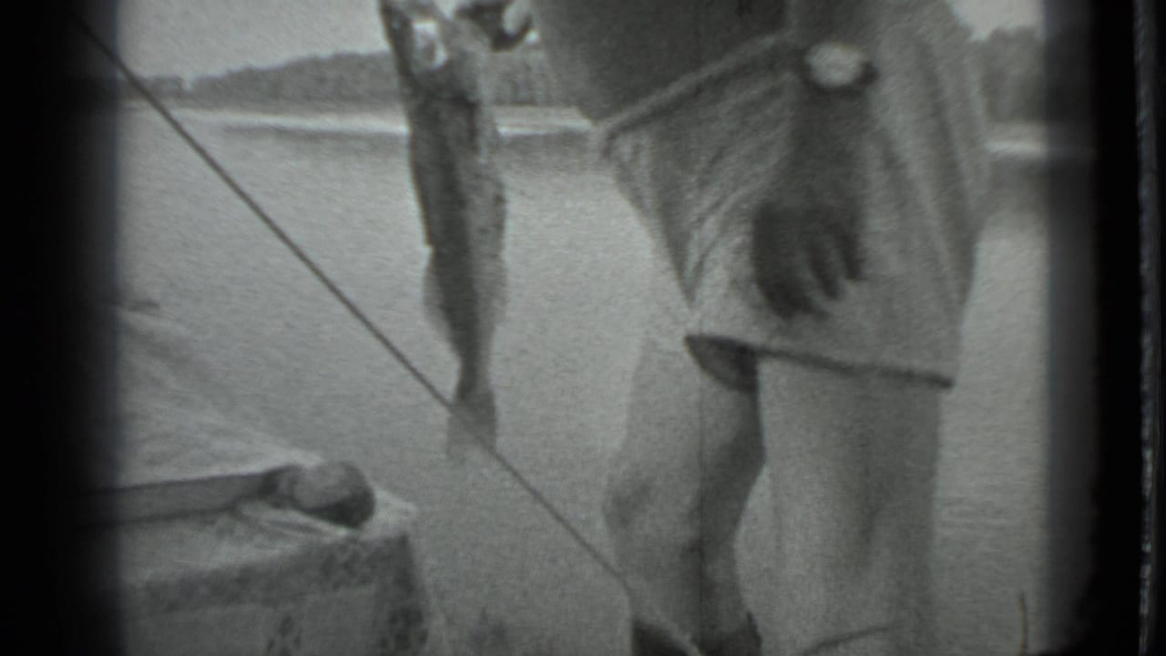 The man stands beside the lake as a large fish hangs vertically from fishing tackle in the 1983 Moscow home-movie scene.