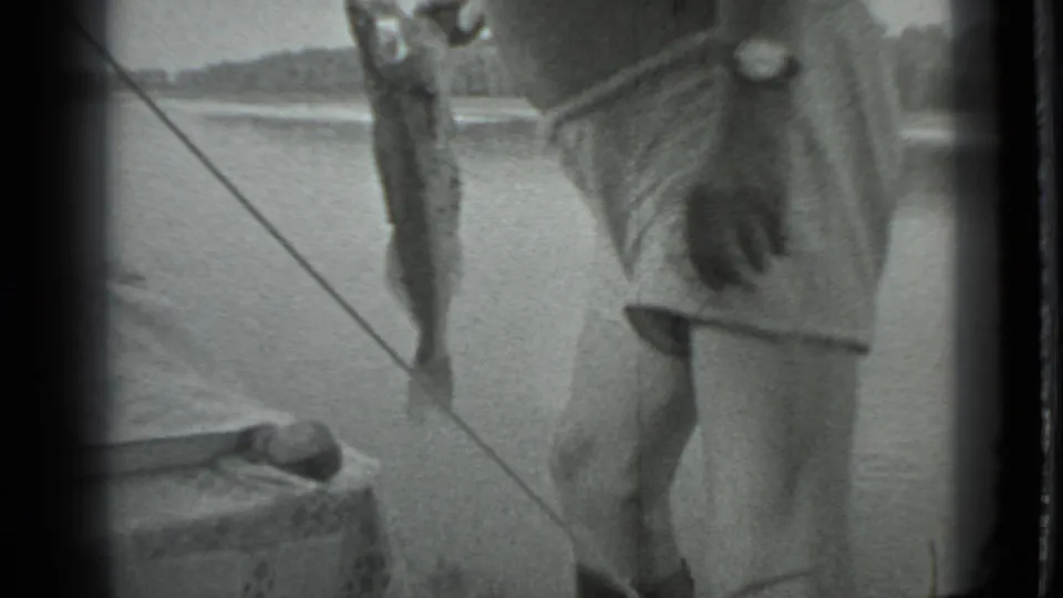 The man stands beside the lake as a large fish hangs vertically from fishing tackle in the 1983 Moscow home-movie scene.