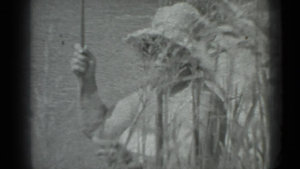A woman in a wide-brimmed hat holds a thin pole or stick among tall grass in Moscow 1983 home-movie footage.