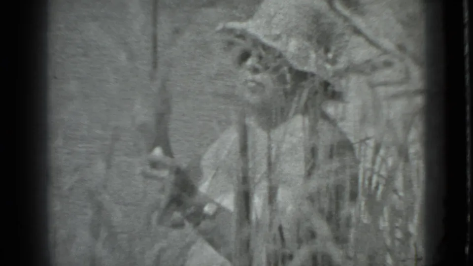 In 1983 Moscow home-movie footage, a person with a wide-brimmed hat stands partly obscured by reeds while holding a fishing rod near the river.