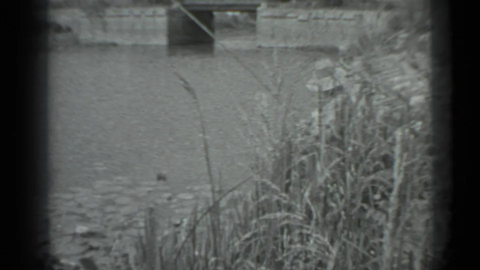 A grainy 8mm view of the Moscow riverbank shows reeds in the foreground, open water, and a low structure beyond the river.