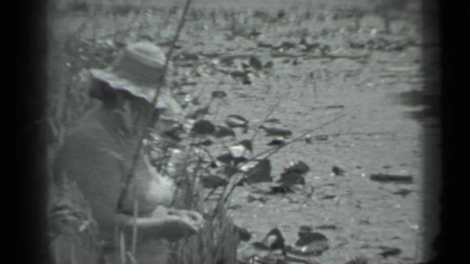 In 1983 Moscow home-movie footage, a mature woman in a straw hat sits near reeds and scattered stones by a shallow riverbank.