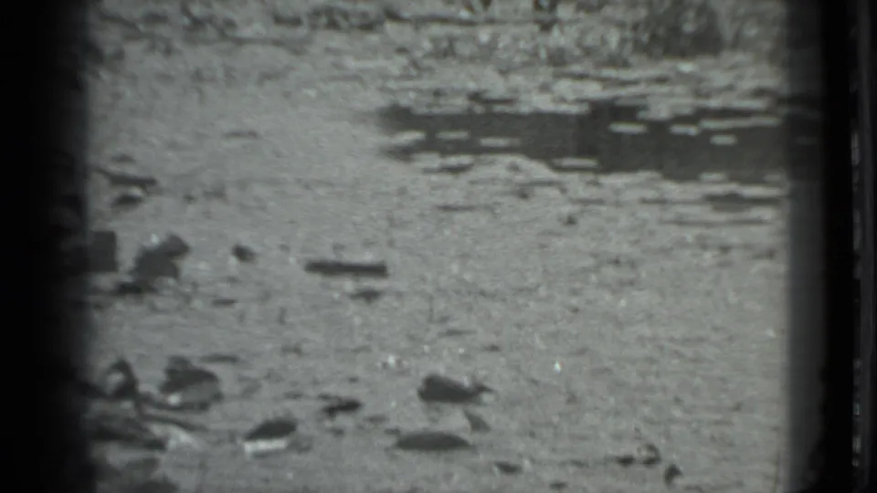 The camera holds on a rocky Moscow riverbank with shallow water and scattered stones, with the woman largely outside the frame.