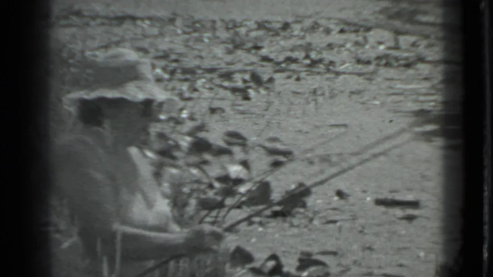 A woman in a light dress and brimmed hat appears to handle a fishing rod or line beside a rocky riverbank in 1983 Moscow.