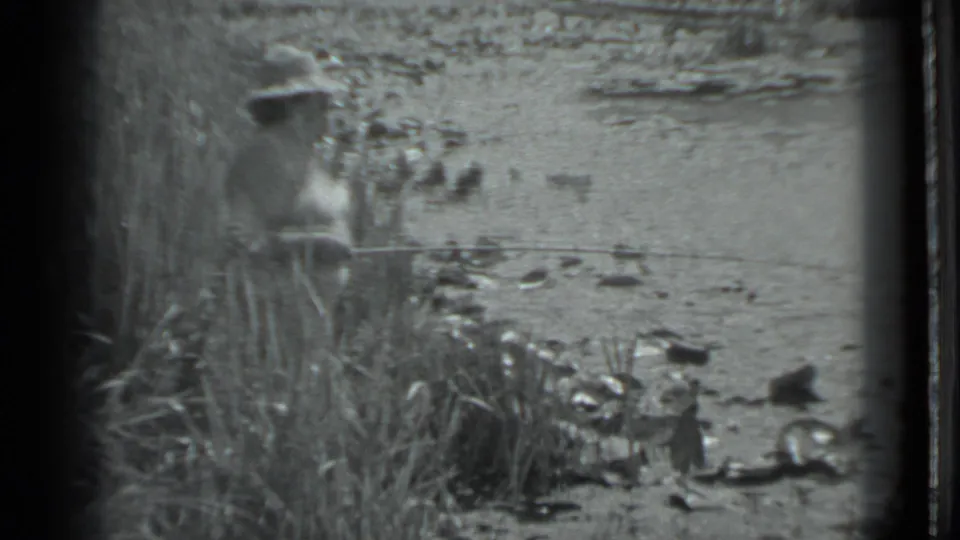 1983 Moscow home-movie frame showing a person in light clothing fishing at a grassy water edge with rocks nearby.