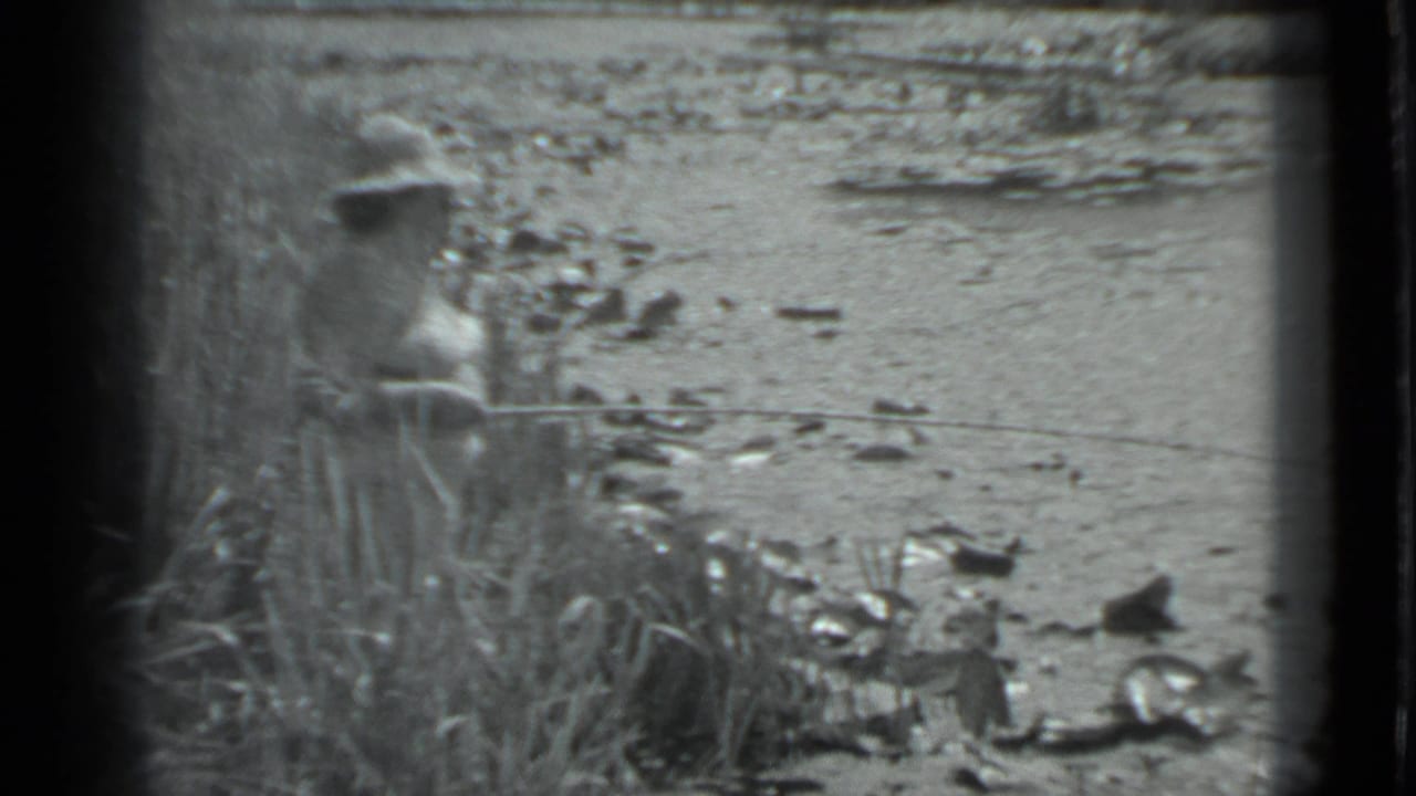 Angler stands partly obscured by grass beside shallow water and rocks.