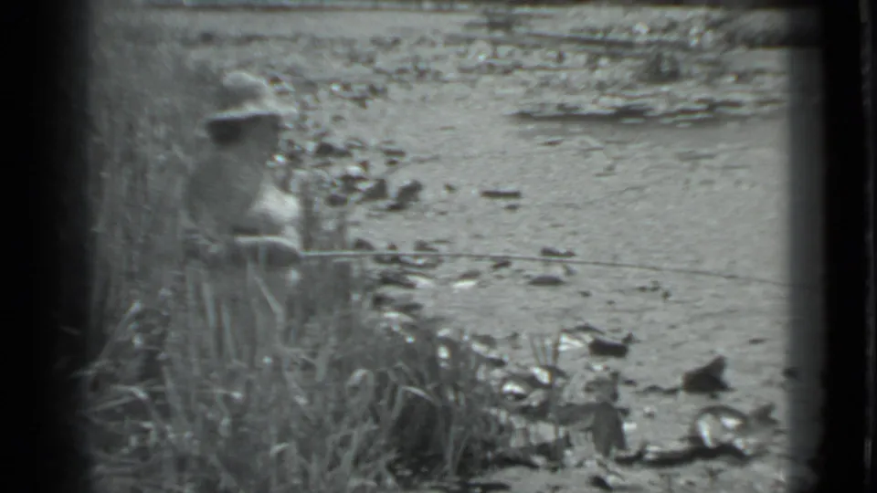 1983 Moscow frame with a person fishing beside reeds or grass, a rod extended across the water, and scattered shoreline rocks.