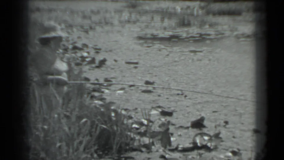 A grainy 8mm frame from Moscow in 1983 shows a person fishing near tall grass and a rocky, plant-covered water edge.
