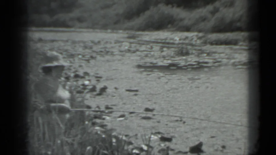 1983 Moscow home-movie still showing a distant angler at a quiet water edge with grass, rocks, and surface plants.