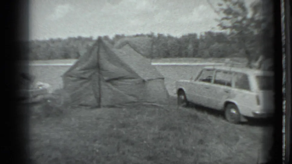 The camera holds on the tent and parked station wagon, with water and dense trees behind the camp area.