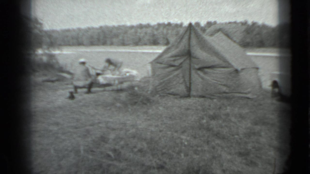 1983 Moscow home-movie frame showing a lakeside tent, seated campers, grass, water, and a wooded far shore.